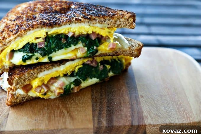 Cheesy Spinach Omelet Melt 3 Close-up of a halved Spinach Omelet Grilled Cheese, showing layers of omelet, spinach, ham, and melted Land O'Lakes cheese, on a cutting board.