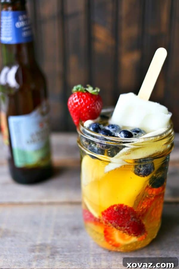 Close-up of Hard Cider Sangria-by-the-Glass, showcasing thinly sliced apples, blueberries, and strawberries mingling in crisp hard cider, with a lime ice pop dissolving and adding flavor and chill.