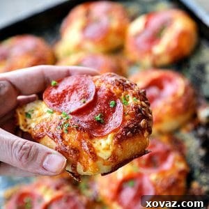 Pepperoni Pizza Bialys Recipe held by hand, with melted cheese, crisp pepperoni, and parsley, against a background of more pizza on an antique cookie sheet.