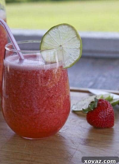 strawberrylimeade2 A close-up of a glass of strawberry limeade, adorned with a fresh lime wedge and half a strawberry on the rim, showcasing its inviting texture and ruby hue.