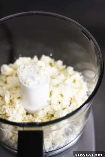 A food processor blending feta cheese, cream cheese, and roasted garlic until smooth.