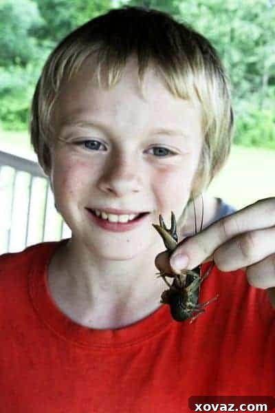 Ty, a smiling young boy, proudly showcases a crawfish he's caught from the creek, his face beaming with the joy and triumph of his outdoor summer fishing adventure with his family.