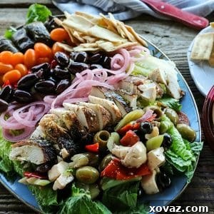 A delectable Greek Salad featuring lemon garlic chicken, a mix of marinated olives, quick pickled red onions, crumbled feta cheese, garlic stuffed Kalamata olives, juicy grape tomatoes, stuffed grape leaves, served with Greek salad dressing, crunchy pita chips, and a rich dollop of hummus. This wholesome salad is perfect for a filling and healthy meal or for convenient meal prep throughout the week.