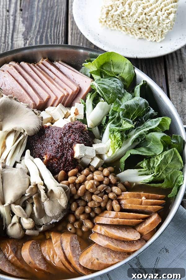Budae Jjigae Korean Spicy Sausage Stew 3 Korean Army Stew or Budae Jjigae in a stainless steel everyday pan, ready to simmer. Bok choy, sliced sausage, sliced spam, mushrooms, baked beans, gochujang seasoning, and ramen noodles on the side on a plate.