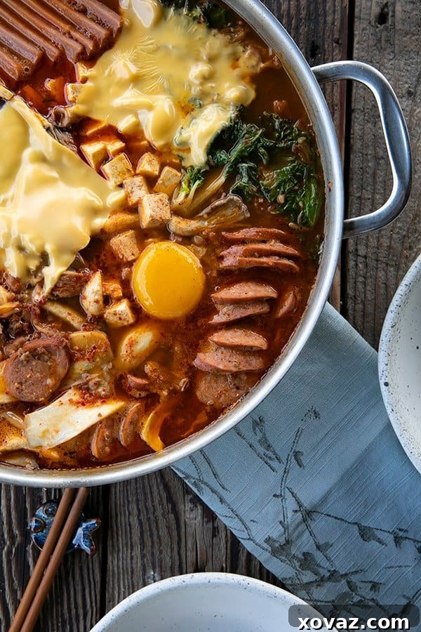 Budae Jjigae Korean Spicy Sausage Stew 5 Korean Army Stew or Budae Jjigae with egg yolks and American cheese in a stainless steel everyday pan. Blue linen on the table with two white bowls and wooden chopsticks.