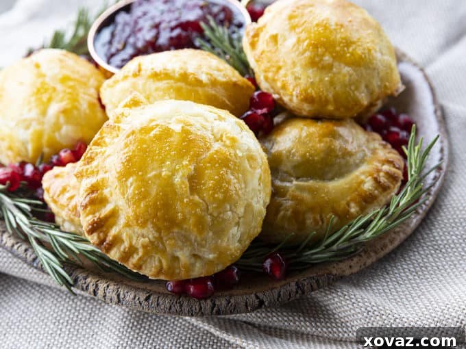 A close-up shot of several bite-sized baked brie en puff pastry appetizers, perfectly golden and flaky, ready to be served. Garnished with fresh herbs and pomegranate seeds.