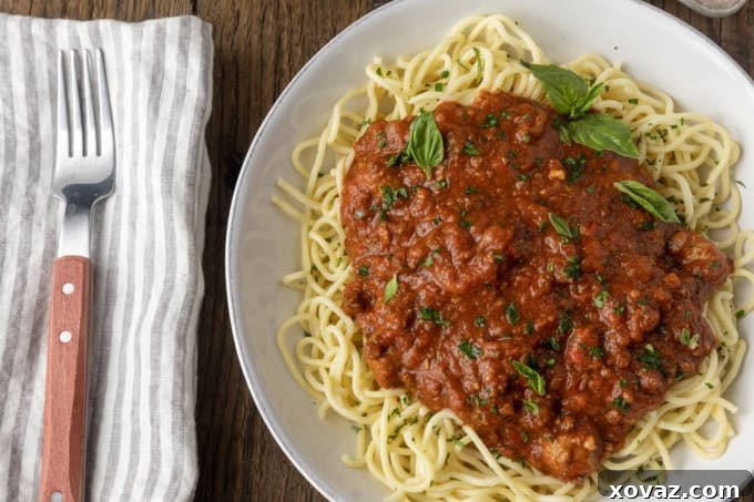 A delectable serving of spaghetti topped generously with homemade crockpot spaghetti sauce and garnished with fresh herbs, alongside a napkin and fork.