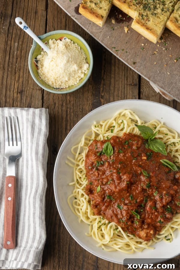 A close-up shot of spaghetti generously topped with slow cooker spaghetti sauce and fresh herbs, with a fork, napkin, and a side of golden garlic bread visible.