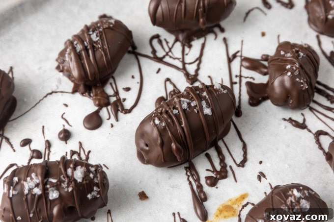 Three almond butter stuffed Medjool dates covered in dark chocolate and sprinkled with sea salt, neatly arranged on a parchment-lined sheet pan.