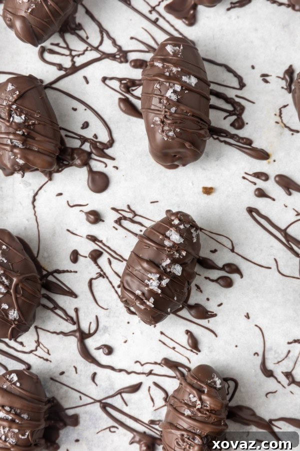 A close-up shot of several chocolate covered almond stuffed dates, lightly dusted with sea salt, cooling on a parchment-lined sheet pan.