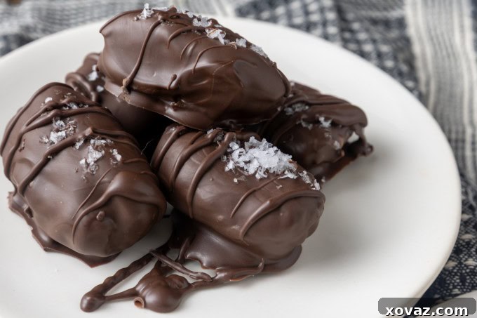 Almond butter stuffed Medjool dates covered in dark chocolate and sprinkled with flaky sea salt, arranged artfully on small white plates with a blue and white linen background.