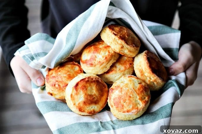 Perfect flaky buttermilk biscuits arranged in a green and white towel-lined bowl, held by a boy in a black long-sleeved shirt on a wooden porch.